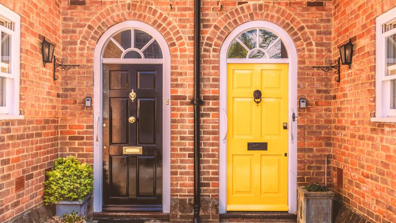 Doors and windows. Two residential front doors, one yellow, one black with a drain pipe down the middle. The walls are red brick there are two side windows and lunette arches over the doors.